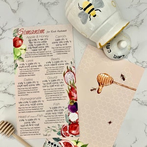 May include: A ceramic honey pot with a bee design, a wooden honey dipper, and two decorative cards with text and illustrations of fruits and honey. The cards have text in Hebrew and English. The theme is Rosh Hashanah.