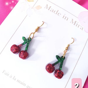 May include: A pair of red and green glitter cherry earrings with gold hooks. The earrings are on a white background with the text "Made in Mira" and "Fait à la main avec amour" in French. The number "2" is written in a pink circle in the bottom right corner of the image.