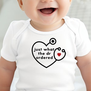 May include: A white baby onesie with a black and red graphic of a heart with a stethoscope and the text "just what the dr ordered".