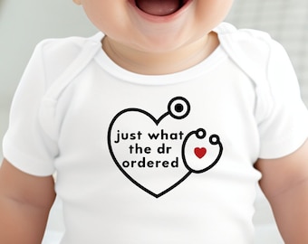 Op de afbeelding: Een witte baby romper met een zwarte en rode afbeelding van een hart met een stethoscoop en de tekst "just what the dr ordered".