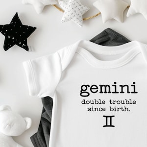 May include: White baby onesie with black text that reads "gemini double trouble since birth. II". The onesie is surrounded by white stars and crescent moons.