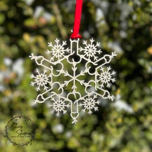 May include: A silver snowflake ornament with a unique design. The snowflake is hanging from a red ribbon against a blurred green background.