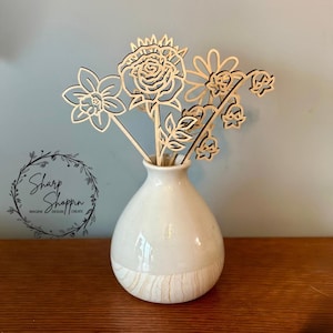 以下が含まれることがあります： 白い陶器の花瓶に、木製の切り抜き花が5つセットされています。花はバラ、水仙、鈴蘭、その他2つの花です。花瓶の底には木目模様があります。