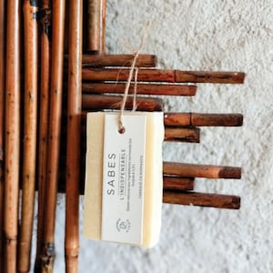 Könnte beinhalten: Eine natürliche Seife mit dem Text "SABES L'INDISPENSABLE Savon naturel aux ingrédients normands bio Surgras à 115% FABRIQUE EN NORMANDIE" auf dem Etikett. Die Seife hängt an einer Schnur und befindet sich vor einem braunen gewebten Hintergrund.