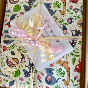 May include: A square gift package wrapped in cream-colored paper featuring a colorful pattern of stylized animals, flowers, and leaves. The package is tied with pale pink and yellow curling ribbons and includes a white card with handwritten text.