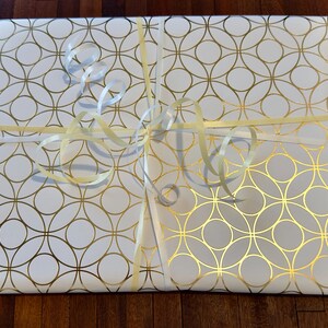 May include: A rectangular package wrapped in white paper featuring a repeating gold geometric circle pattern. The package is tied with a thin, pale yellow ribbon that is knotted in the center and includes several curled ribbon ends on top.