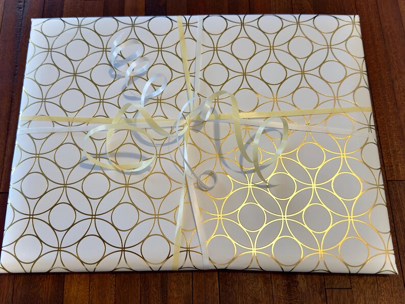 May include: A rectangular package wrapped in white paper featuring a repeating gold geometric circle pattern. The package is tied with a thin, pale yellow ribbon that is knotted in the center and includes several curled ribbon ends on top.