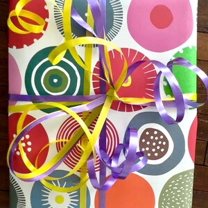 May include: A rectangular gift wrapped in white paper featuring a colorful pattern of circles in shades of red, pink, orange, green, and blue, decorated with intersecting bright yellow and purple curling ribbons tied in a central knot.