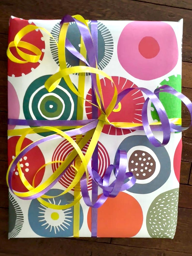 May include: A rectangular gift wrapped in white paper featuring a colorful pattern of circles in shades of red, pink, orange, green, and blue, decorated with intersecting bright yellow and purple curling ribbons tied in a central knot.