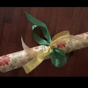 May include: A cylindrical object wrapped in cream-colored paper featuring a repeating pattern of red poinsettias, green pine needles, and small brown pinecones. The package is tied with a solid green satin ribbon and a sheer gold ribbon.