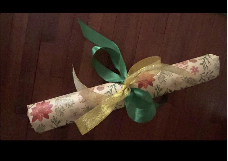 May include: A cylindrical object wrapped in cream-colored paper featuring a repeating pattern of red poinsettias, green pine needles, and small brown pinecones. The package is tied with a solid green satin ribbon and a sheer gold ribbon.