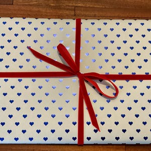 May include: A rectangular gift box wrapped in white paper featuring a repeating pattern of small, metallic blue hearts. The package is tied with a thin, vibrant red velvet ribbon that is knotted into a bow at the center of the top surface.