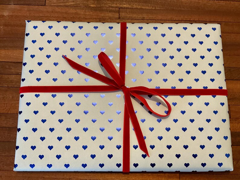 May include: A rectangular gift box wrapped in white paper featuring a repeating pattern of small, metallic blue hearts. The package is tied with a thin, vibrant red velvet ribbon that is knotted into a bow at the center of the top surface.