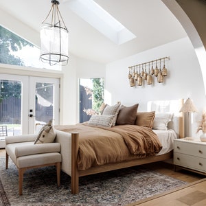 May include: A bedroom with a large bed with a brown duvet and white pillows. The bed is next to a window with a view of a backyard. There is a white nightstand with a lamp on it. The room has a white ceiling with a skylight and a large chandelier.
