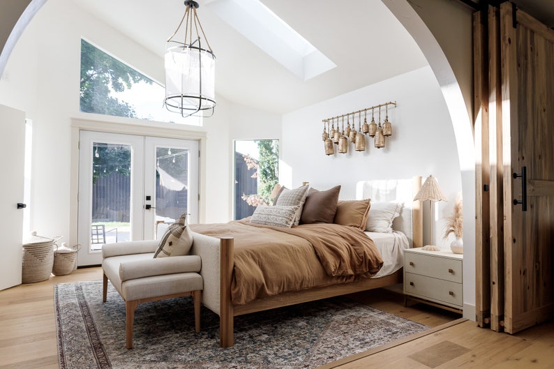 May include: A bedroom with a large bed with a brown duvet and white pillows. The bed is next to a window with a view of a backyard. There is a white nightstand with a lamp on it. The room has a white ceiling with a skylight and a large chandelier.