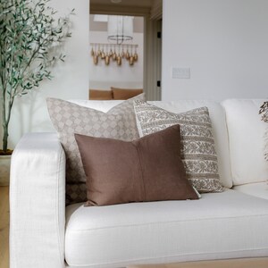 May include: A white couch with brown and beige patterned throw pillows. The couch is in a living room with a potted olive tree and a wooden coffee table.