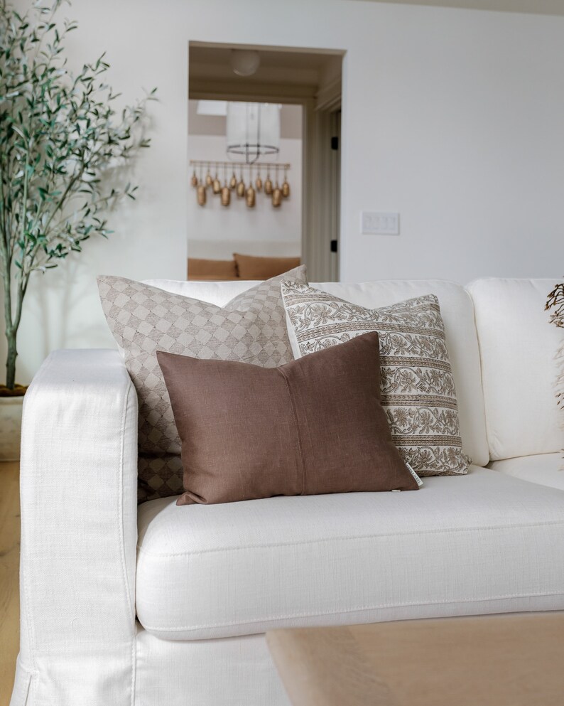 May include: A white couch with brown and beige patterned throw pillows. The couch is in a living room with a potted olive tree and a wooden coffee table.