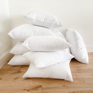 May include: A stack of eight white down pillows of various sizes, arranged on a wooden floor.
