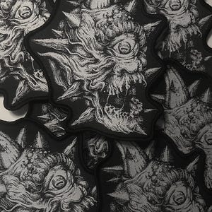 May include: A collection of black and white patches featuring a detailed illustration of a monster with a single eye, sharp teeth, and spiky protrusions. The patches have a star-like shape.