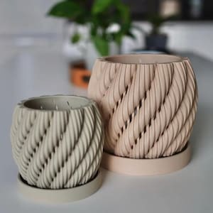 May include: Two cylindrical planters with a spiraling rope-like texture on the exterior. The smaller planter is light sage green, and the larger planter is a soft peach color. Each planter sits on a matching circular saucer base.