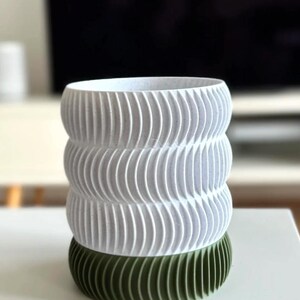 May include: A white and green decorative vase with a unique, textured design. The vase features a stacked, cylindrical shape with a ribbed pattern. The base is a dark green color, while the upper portion is white.