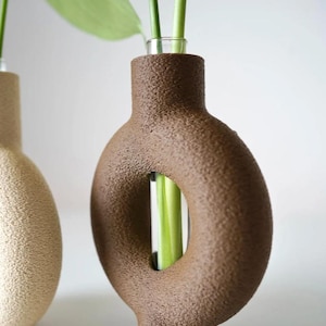 May include: A brown, textured ceramic vase with a circular design and a small glass tube for holding stems. The vase has a unique, modern shape and is filled with green stems and leaves. Another vase is visible to the left.