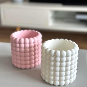 May include: Two cylindrical planters with a textured, bubble-like design. One is light pink, the other is white. The planters are on a light gray surface, with a blurred white cabinet in the background. These decorative planters are suitable for small plants or as desk organizers.