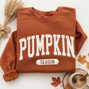 May include: A rust-colored crewneck sweatshirt featuring the white text PUMPKIN in a large arched font and the word SEASON in a smaller white rectangular patch below. The garment has long sleeves with ribbed cuffs and a soft interior lining.