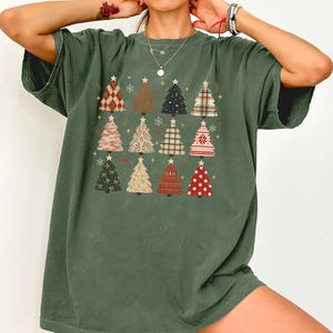 May include: A dark olive green short-sleeve t-shirt featuring a graphic print of twelve stylized Christmas trees arranged in a grid. Each tree displays a unique pattern, including plaid, polka dots, camouflage, and floral designs, topped with a star.