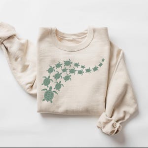 May include: A beige sweatshirt featuring a design of green sea turtles swimming in a curved line. The turtles vary in size, creating a sense of movement. The sweatshirt has a crew neck and long sleeves with rolled-up cuffs.