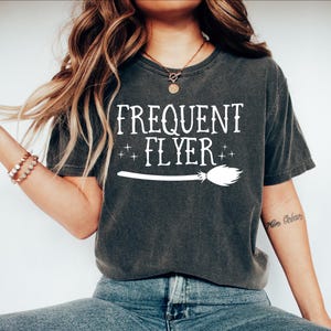 May include: A charcoal gray short-sleeve t-shirt featuring the white text "FREQUENT FLYER" printed above a graphic of a witch's broomstick and small star accents. The shirt has a crew neckline and a relaxed fit, made of a washed-out fabric texture.