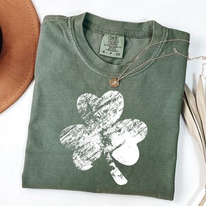 May include: A green t-shirt featuring a white distressed shamrock design. The shirt has a crew neck and is made from a soft, comfortable fabric. The brand name "COMFORT COLORS" is visible on the tag.
