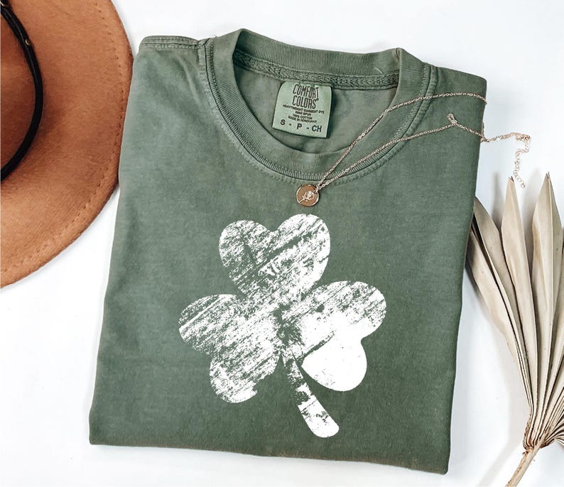May include: A green t-shirt featuring a white distressed shamrock design. The shirt has a crew neck and is made from a soft, comfortable fabric. The brand name "COMFORT COLORS" is visible on the tag.
