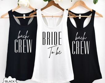 May include: Three tank tops with black and white text. The center tank top is white with black text that reads "BRIDE To Be". The tank tops on either side are black with white text that reads "bach CREW".