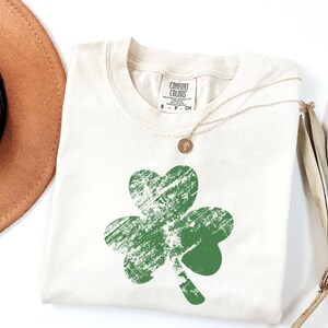May include: A white t-shirt featuring a green shamrock design. The shamrock has a distressed, vintage look. The shirt has a tag that says "Comfort Colors".