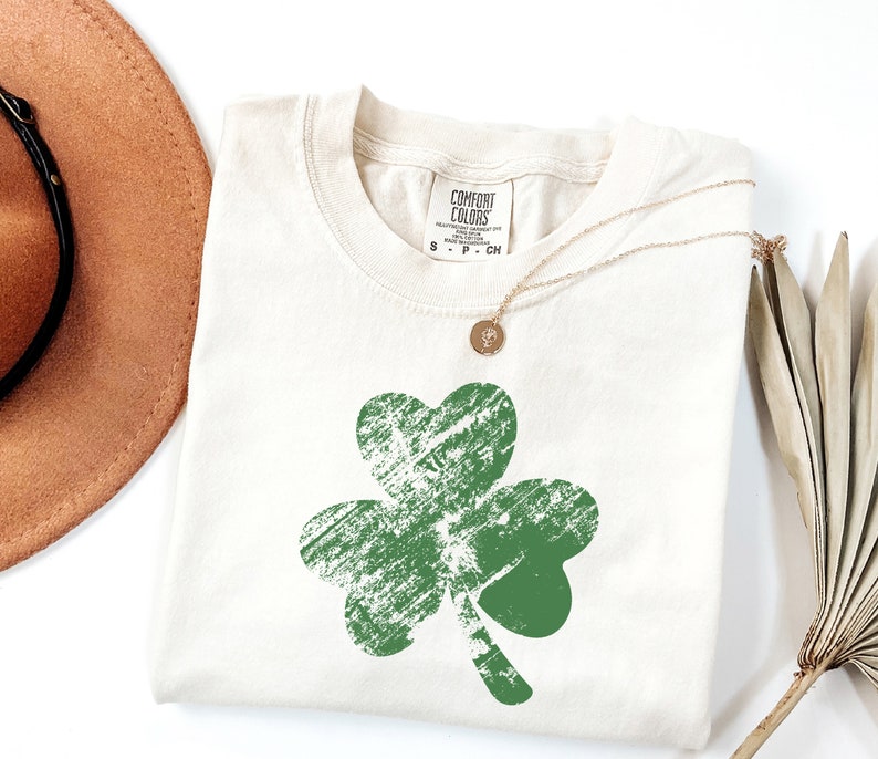 May include: A white t-shirt featuring a green shamrock design. The shamrock has a distressed, vintage look. The shirt has a tag that says "Comfort Colors".