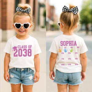May include: A child wearing a white t-shirt with the text "CLASS OF 2038" in purple. The child is also wearing denim shorts, heart-shaped sunglasses, and a black and white bow. The back of the shirt has the name "SOPHIA" and handprints.