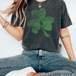 May include: A dark grey t-shirt with a distressed green shamrock graphic. The shirt has a relaxed fit and a crew neck.
