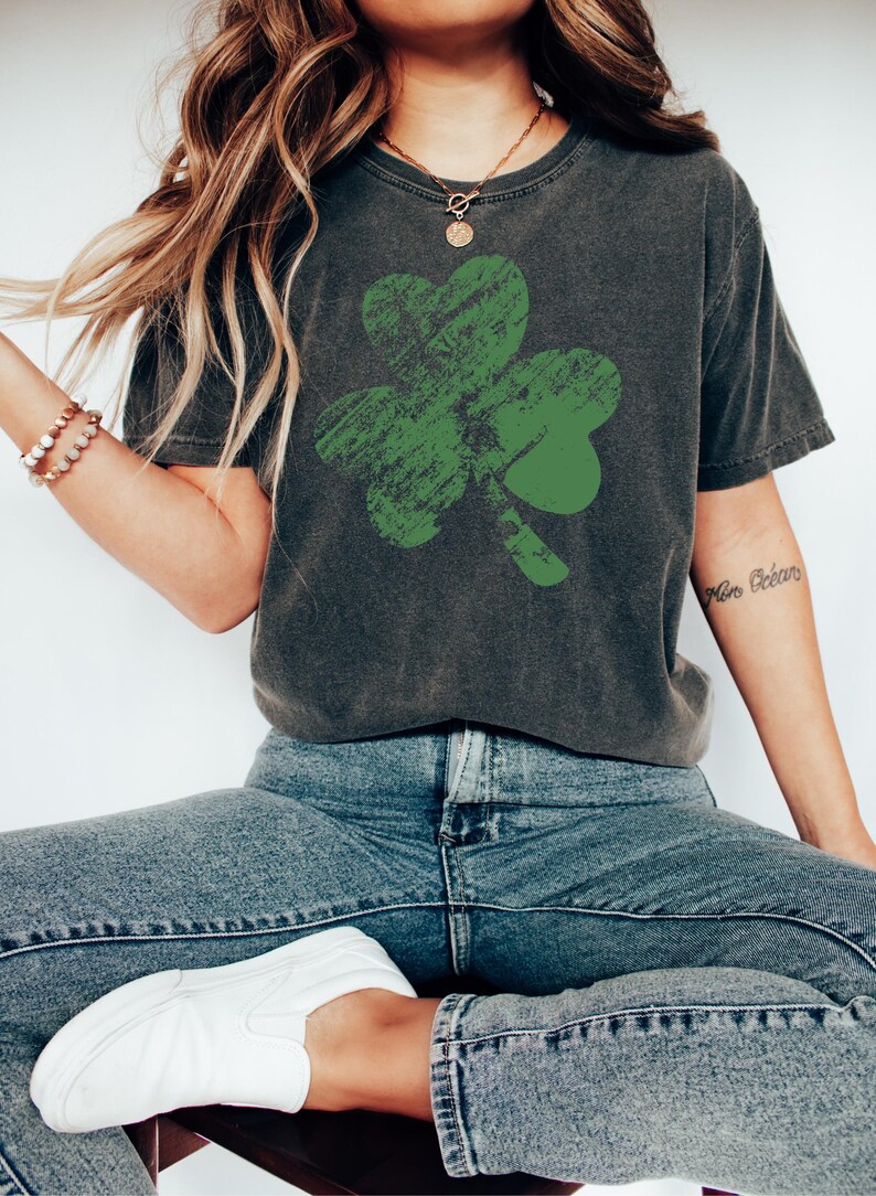 May include: A dark grey t-shirt with a distressed green shamrock graphic. The shirt has a relaxed fit and a crew neck.