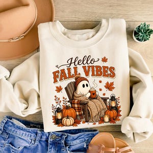 May include: A cream-colored crewneck sweatshirt featuring a graphic of a ghost sitting in a wooden chair, holding a mug, surrounded by pumpkins, lanterns, and maple leaves. The text "Hello FALL VIBES" is printed in brown and orange above the ghost.