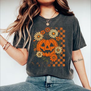 May include: Dark gray t-shirt featuring a vintage-style Halloween design. The graphic includes an orange jack-o'-lantern surrounded by daisies and other orange flowers, with a checkered pattern on the right side. The shirt has a distressed look.