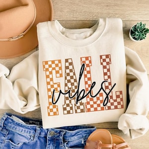 May include: Cream-colored sweatshirt with the words "Fall Vibes" in a checkered pattern of orange and brown. The sweatshirt is paired with blue denim shorts, a brown hat, and sandals. A small succulent plant is also visible.