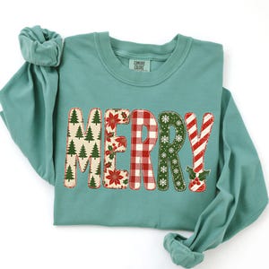 May include: A teal long-sleeve crewneck sweatshirt featuring the word MERRY in large, patterned letters. Each letter has a unique design: pine trees, poinsettias, red and white plaid, white snowflakes on green, and red and white stripes.