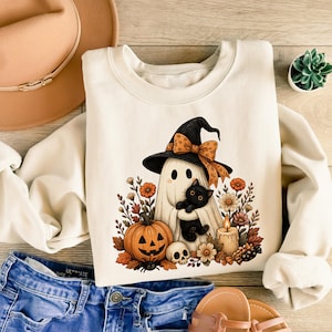 May include: A cream-colored crewneck sweatshirt featuring a graphic of a ghost wearing a witch hat and holding a black cat. The design includes a jack-o'-lantern, a skull, a lit candle, and autumn flowers. The sweatshirt is displayed with a hat.