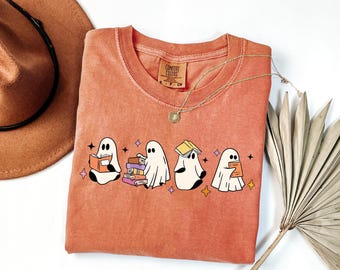 Può includere: Una t-shirt color terracotta con una grafica di quattro fantasmi bianchi che interagiscono con dei libri. Un fantasma legge, uno impila libri, uno ne tiene uno in equilibrio sulla testa e uno ne tiene uno in mano. Piccole stelle colorate sono sparse intorno.