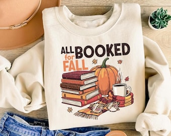 Peut inclure: Sweat-shirt crème avec le texte "ALL BOOKED for FALL" et un graphisme de livres, une citrouille, une tasse à café et des lunettes. Le sweat-shirt est associé à un jean bleu, un chapeau marron et des sandales.