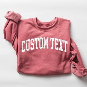 May include: A red-pink sweatshirt with the text "CUSTOM TEXT" in white block letters. The sleeves are cuffed, and the crew neck is visible. The sweatshirt is made of a soft material.