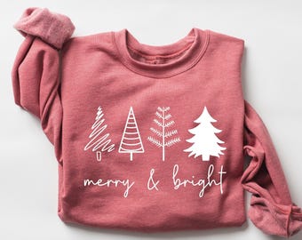 May include: A dusty rose long-sleeve sweatshirt featuring four distinct white graphic illustrations of pine trees arranged in a row. Below the trees, the text "merry & bright" is printed in a white, cursive, handwritten-style font.