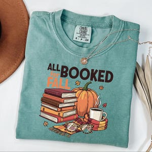 May include: Teal-colored t-shirt with the text "ALL BOOKED FOR FALL" and a graphic of books, a pumpkin, a coffee cup, and glasses. A silver necklace with a pendant is draped across the shirt.