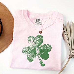 May include: Pink t-shirt with a green shamrock graphic. The shamrock has a distressed look and is centred on the shirt. The shirt has a crew neck and short sleeves.